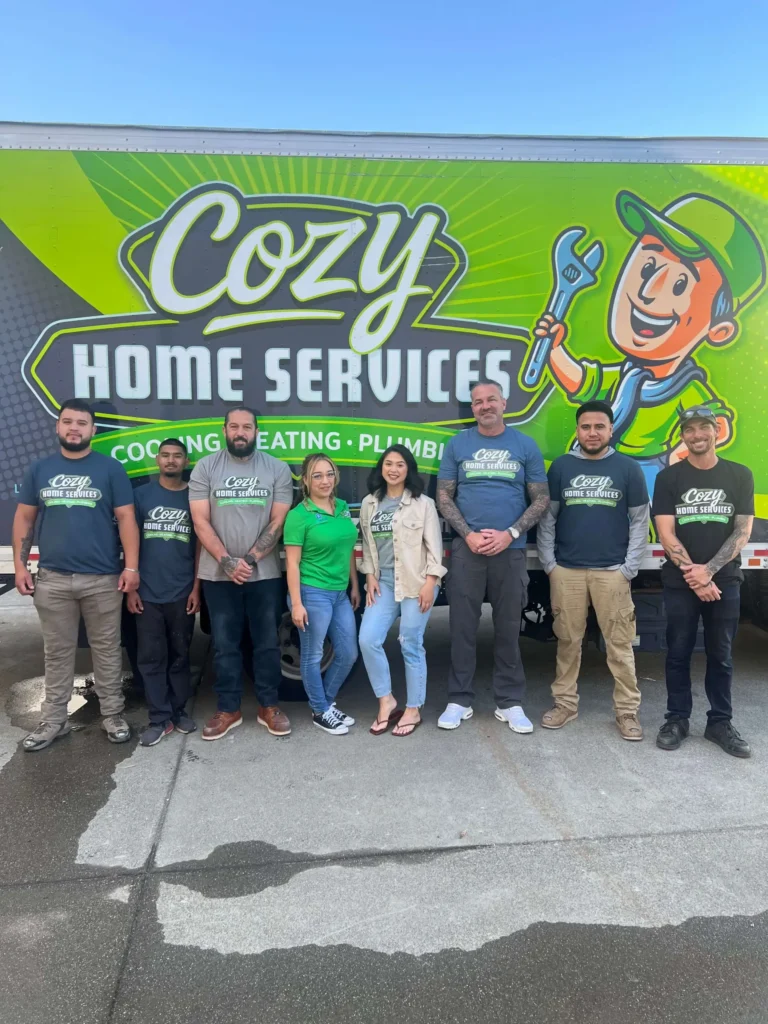 Eight people stand in front of a green Cozy Home Services truck with a cartoon mascot and bold logo, posing for a group photo on a sunny day—a perfect addition to the company’s Photo Gallery.