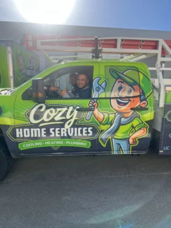 Two men sit inside a bright green Cozy Home Services van featuring a cartoon mascot and text advertising cooling, heating, and plumbing services.